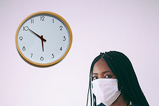 Female African American patient wearing purple mask waiting for doctor next to a clock on a purple wall.