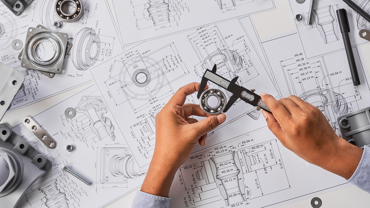  Mechanical engineer measuring ball bearings with caliper tool on table with multiple design drawings.